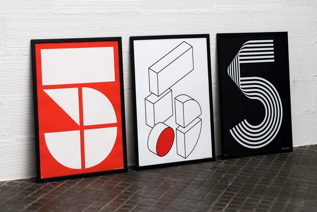 Three framed graphic prints in red, black and white leaning against a white brick wall on a dark floor
